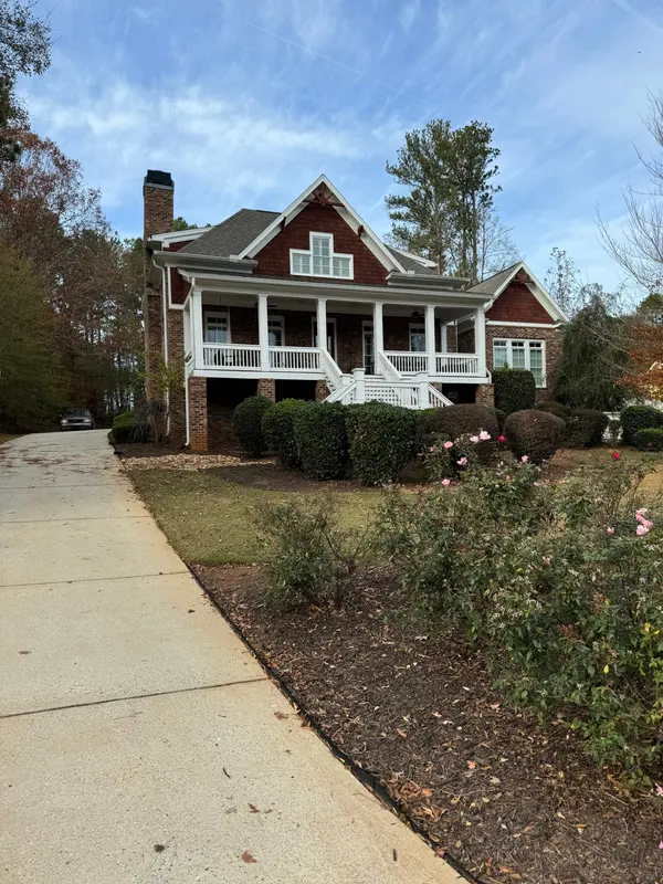 Large two-story home with a red siding upper story and white wraparound porch seen from the drive