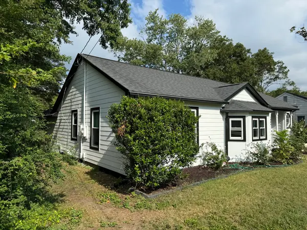 Corner of a white painted cottage with black trim and a bay window behind a shrub