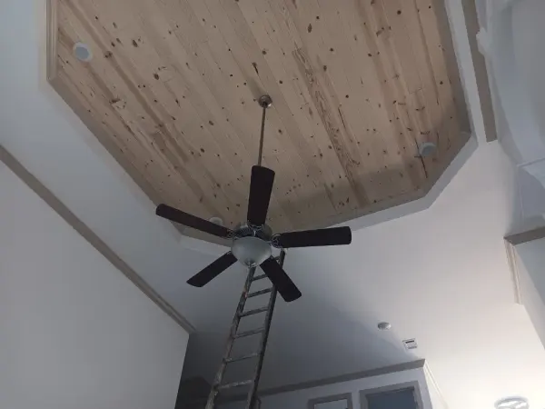 Raw pine plank tray ceiling with a fan and a ladder below, white walls